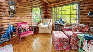 A log cabin interior with a wooden floor, a red floral sofa, a beige armchair, a wooden cabinet, and a clock on the wall. - Unplug! Lake Toxaway Cabin w/ Dock Access (Lake Toxaway)
