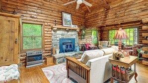 A log cabin interior with a stone fireplace, wooden furniture, and a ceiling fan. - Unplug! Lake Toxaway Cabin w/ Dock Access (Lake Toxaway)