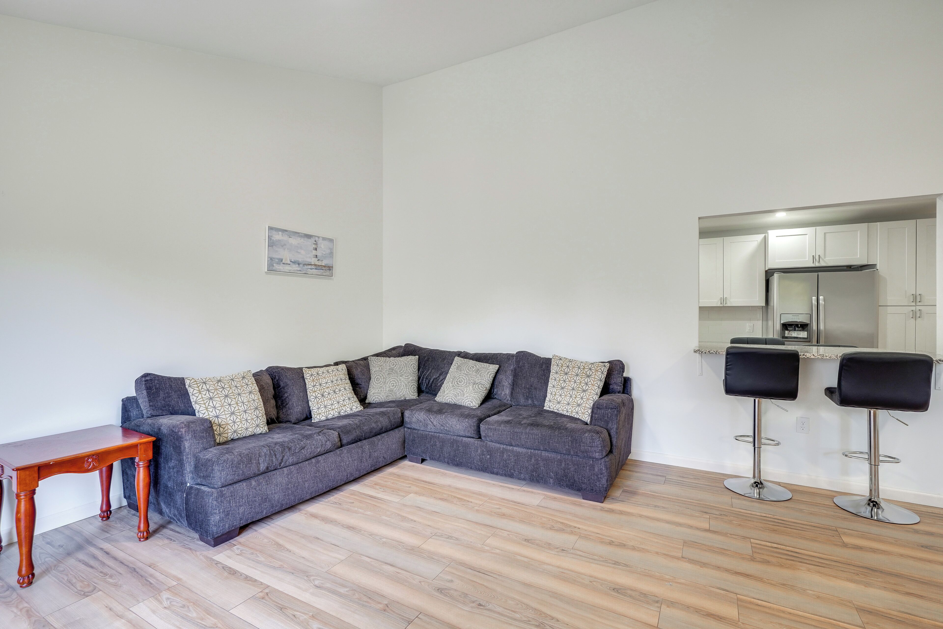 Ein Wohnzimmer mit grauem Ecksofa, zwei schwarzen Barhocker, einem Holztisch und einer Küchenzeile mit weißen Schränken und einem Edelstahlkühlschrank.