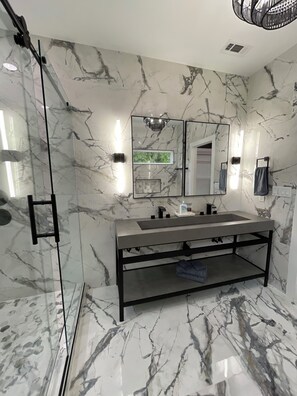 A modern bathroom with a glass shower enclosure, a marble-tiled wall, a large mirror, and a built-in sink.