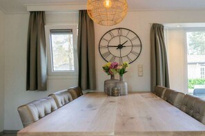 A dining room with a long table, cushioned chairs, a large wall clock, and a hanging light fixture.