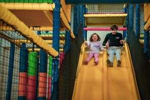 Two children sliding down a yellow slide in a play area with netted sides and colorful padded structures. - Hackfort l"etage Sauna 6 (Lochem)