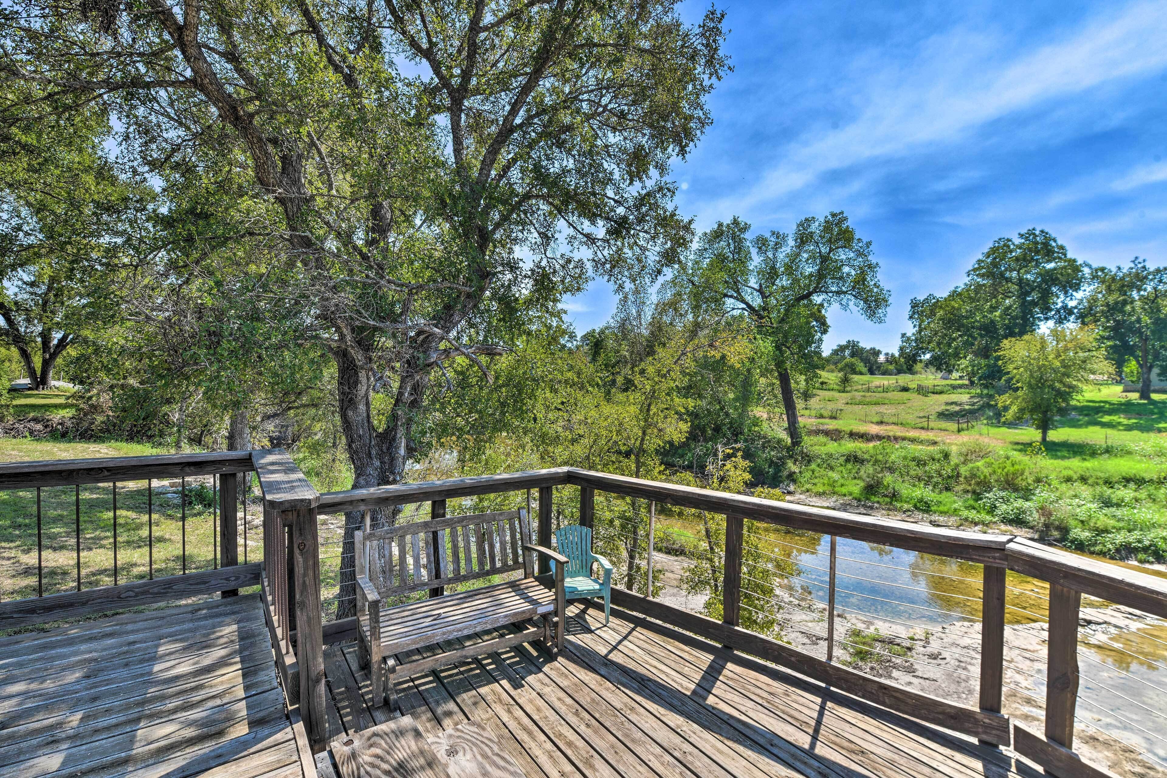 Terraza de madera con banco y silla, vista a un río y follaje.