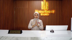 A reception desk with a 'RESEPSIONIS' sign, a woman standing with hands in a prayer position, and a 'Nimo Resort' sign in the background. - Nimo Resort Ciater (Ciater)