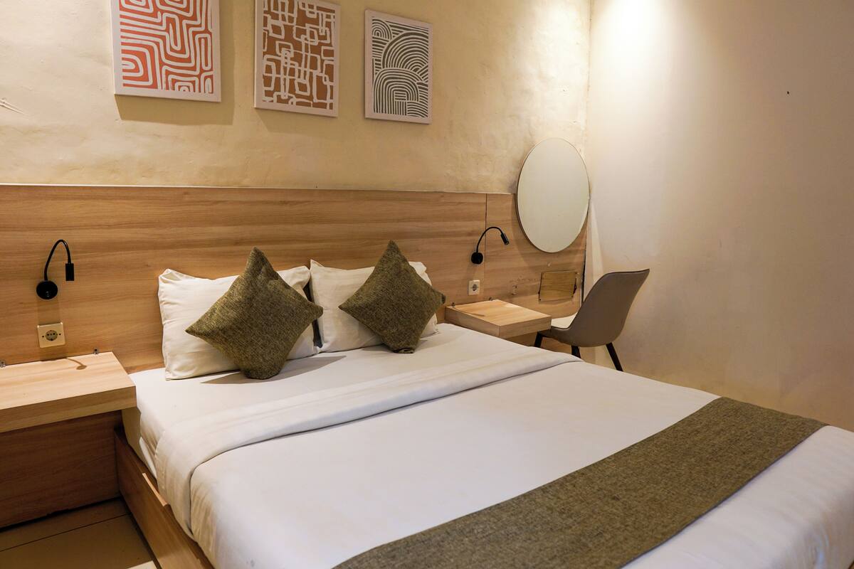 A hotel room with a wooden headboard, two bedside tables, a round mirror, and a bed with white bedding and two decorative pillows.