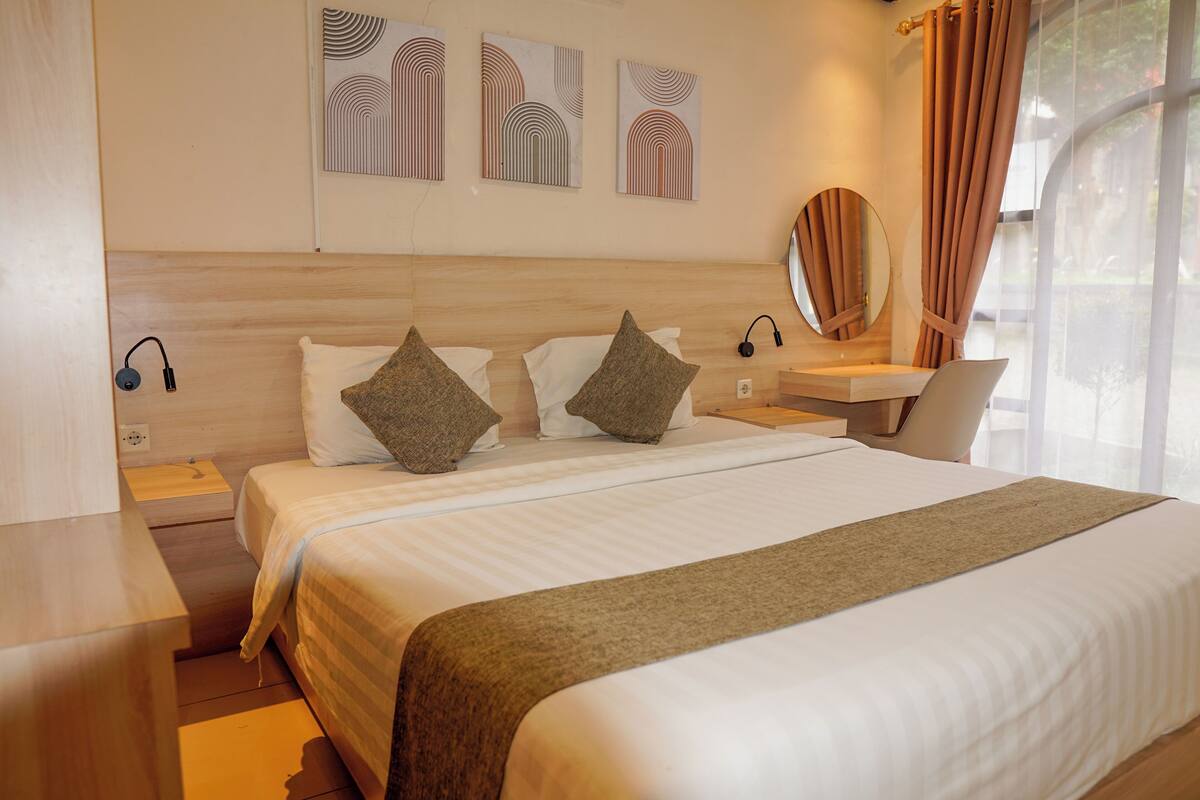 A modern hotel room with a large bed, a bedside table with a lamp, a laptop, and a mirror.