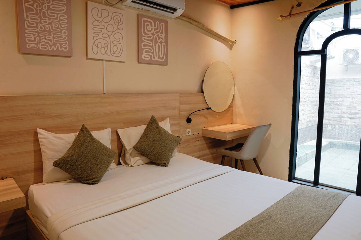 A hotel room with a bed, a desk, a chair, a large window, and artwork on the wall.