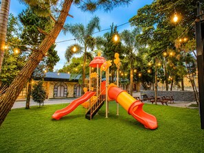 A playground with slides and climbing structures, surrounded by trees and string lights.