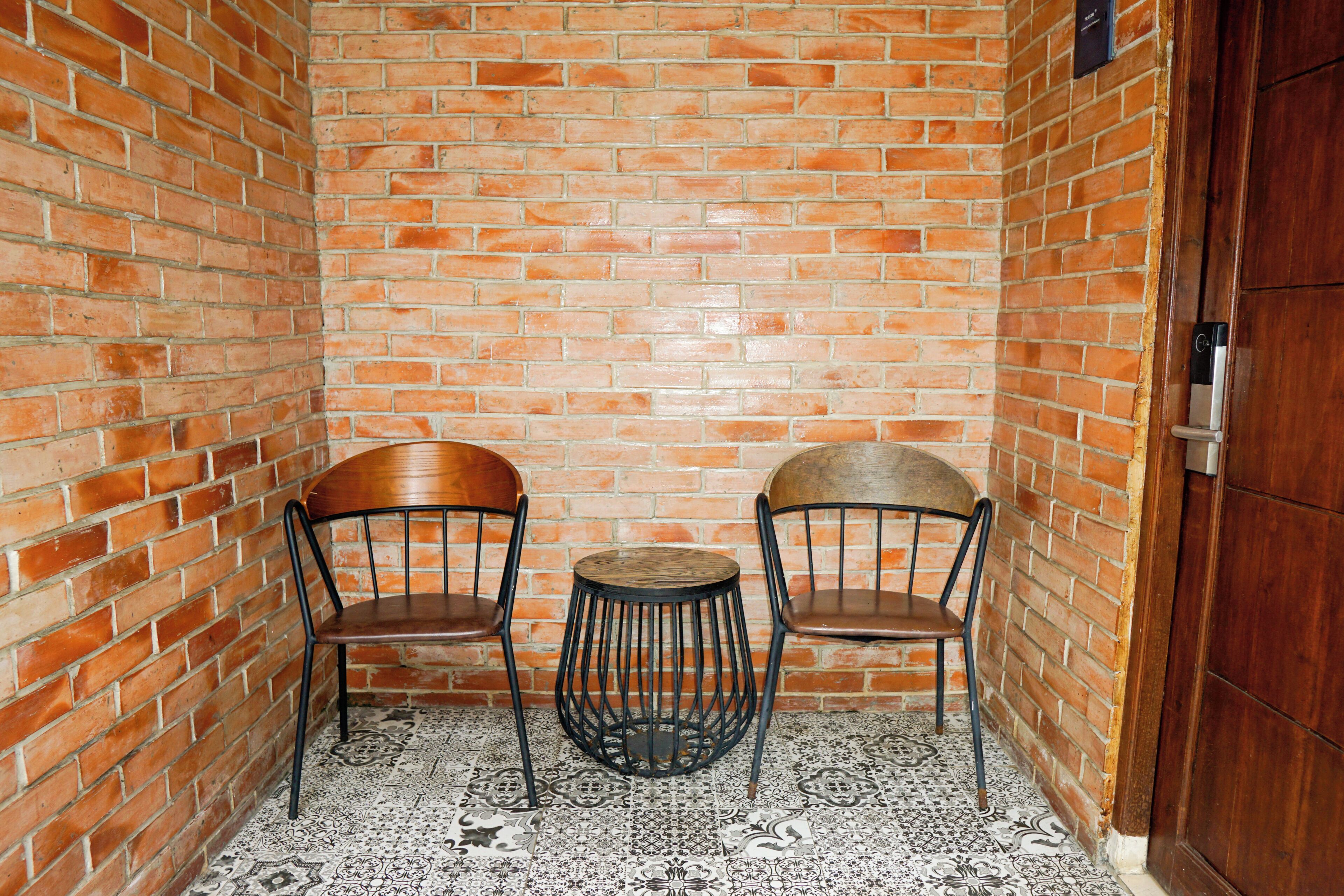 A small, enclosed seating area with two wooden chairs and a round table, set against a brick wall with a patterned floor.
