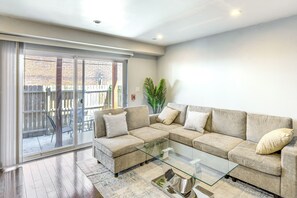 Ein Wohnzimmer mit einem beigefarbenen Ecksofa, einem Glastisch und einer Glasschiebetür, die auf eine Terrasse im Freien führt.