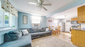 Ein Wohnzimmer mit grauem Ecksofa, Deckenventilator und einer Küchenzeile mit Holzschränken und Geräten.