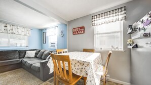 Ein Wohnzimmer mit Ecksofa, einem Esstisch mit Stühlen, einer Küchenzeile mit Kühlschrank und einem Fenster mit Jalousien.