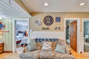 A cozy living room with a large clock, a couch with patterned cushions, and a wall with framed photos and a 'beach' sign. - George Lake Escape! Charming 1950s Cabin (Lupton)