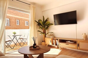 Una sala de estar con una mesa redonda, un televisor, una planta y un balcón con mesa y sillas.