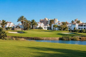 Un campo de golf con una masa de agua, palmeras y edificios al fondo.