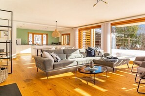 Ein modernes Wohnzimmer mit grauem Sofa, rundem Couchtisch und einem großen Fenster mit Jalousien.