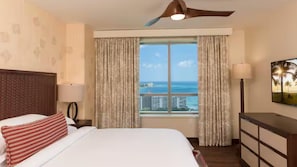 Quarto de hotel com cama, ventilador de teto, janela com cortinas, luminária e vista para o mar.