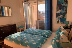 A bedroom with a bed, a dresser, a chair, a painting, and a view through a glass door. - Holiday apartment with 1 bedroom (Garmisch-Partenkirchen)