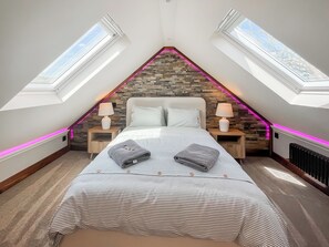 Ein Schlafzimmer mit Schrägdecke, Dachfenstern, einem Bett mit gestreifter Bettwäsche, zwei Nachttischen mit Lampen und einer Wand aus Naturstein.
