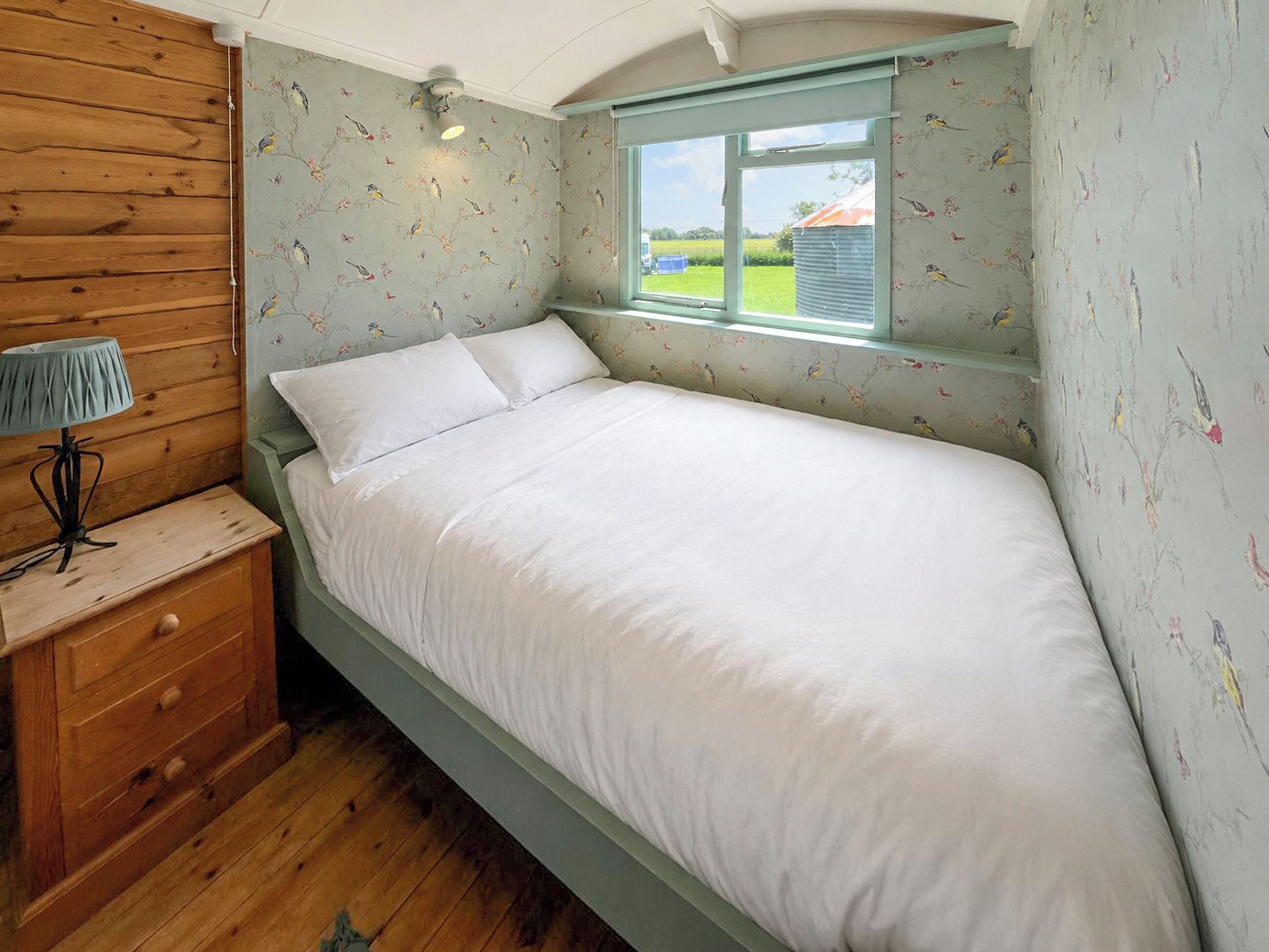 A single bed with white bedding, a wooden nightstand with drawers, a lamp, and a window with a view of the outside.