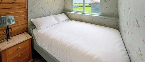 A single bed with white bedding, a wooden nightstand with drawers, a lamp, and a window with a view of the outside.