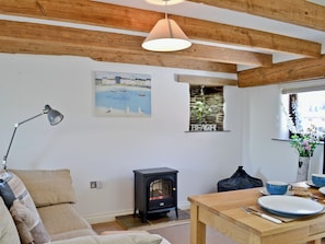 Ein gemütliches Zimmer mit Holzboden, einem Bild einer Strandansicht, einem kleinen Fenster mit der Aufschrift 'BEACH' sowie einem Tisch mit Teller und Tasse und einem schwarzen Kaminofen.