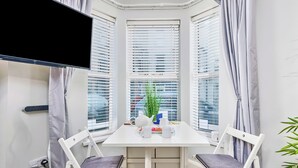 Une pièce d’angle avec une table blanche, deux chaises, un téléviseur et une fenêtre avec des stores.