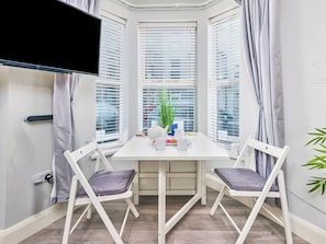 Ein Eckzimmer mit weißem Tisch, zwei Stühlen, einem Fernseher und einem Fenster mit Jalousien.