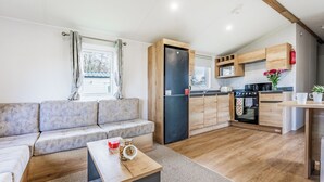 Een knus woongedeelte met een bank, een klein bijzettafeltje, een keuken met houten kastjes en apparatuur, en een raam met gordijnen.