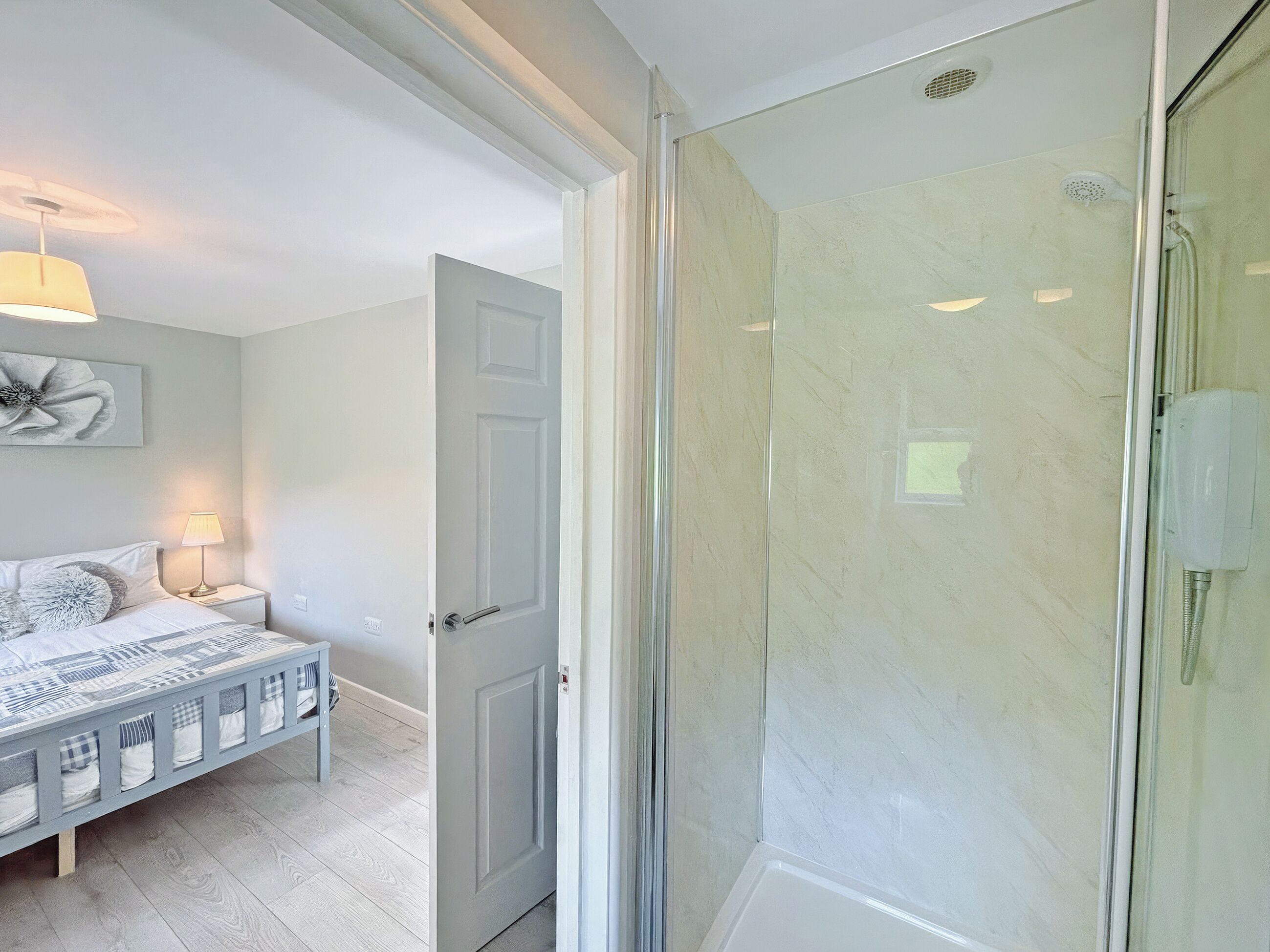 Un dormitorio con cama, mesita de noche, lámpara y puerta que da a una zona de ducha con mampara de vidrio y cabezal de ducha.
