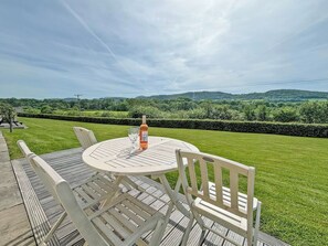 A round outdoor table with chairs, a bottle of wine, and glasses on a deck overlooking a well-manicured lawn and distant hills.