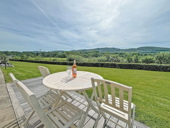 A round outdoor table with chairs, a bottle of wine, and glasses on a deck overlooking a well-manicured lawn and distant hills.