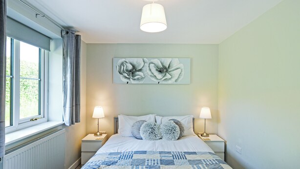 Een slaapkamer met een bed, twee bedlampen, een raam met gordijnen, een radiator en een bloemschilderij aan de muur.