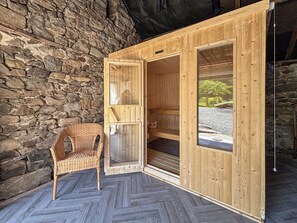 A wooden sauna room with a stone wall, a wicker chair, and a view of a gravel path. - 2 bedroom accommodation in Rhayader and the Elan Valley (Rhayader and the Elan Valley)