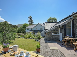 Ein Haus mit Steinfassade, ein Garten mit einem Holztisch, der für ein Kaffeetrinken gedeckt ist, und Blick auf die umliegende Landschaft.