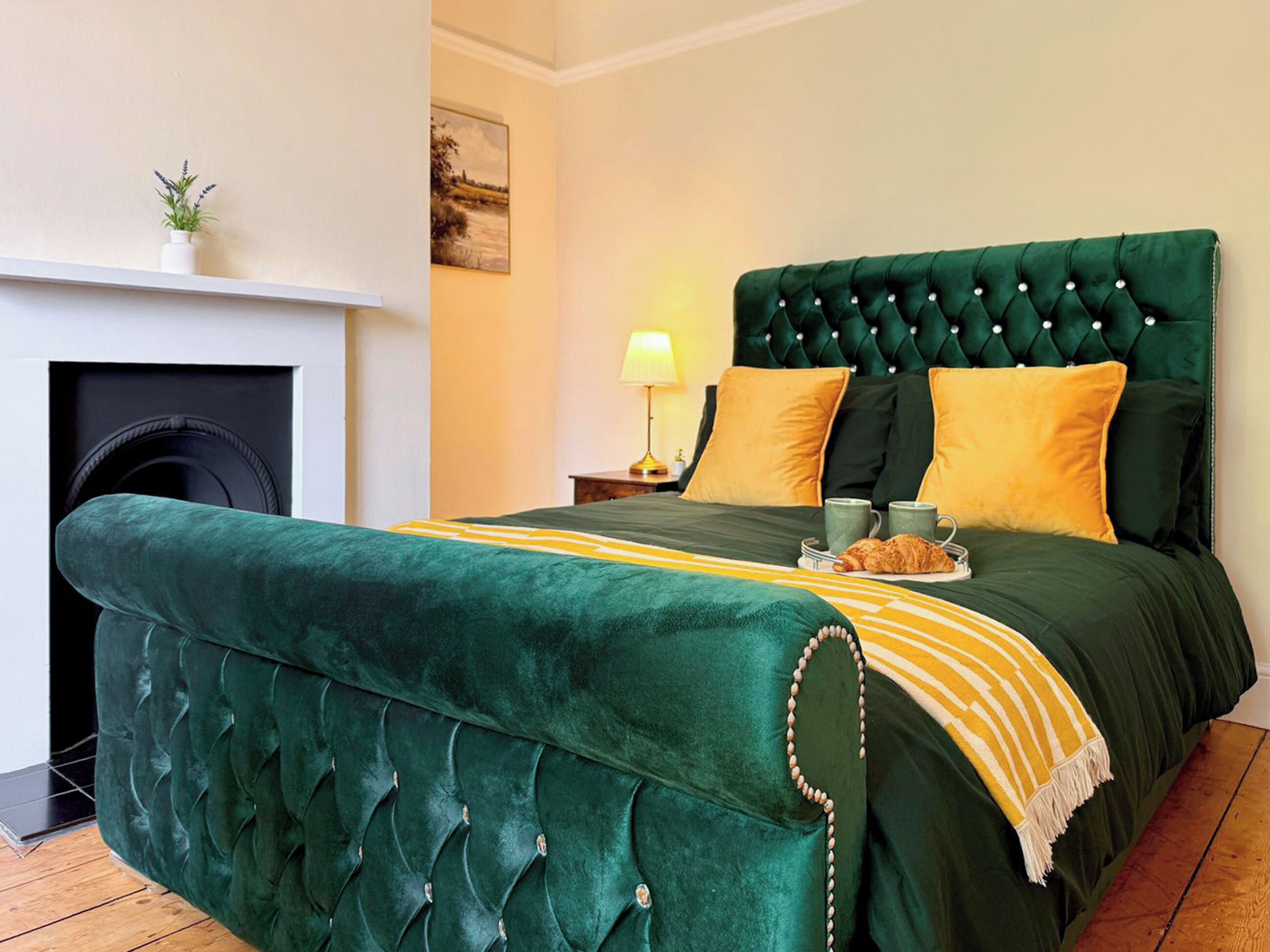 Une chambre à coucher avec un lit recouvert d’un tissu vert, une cheminée, une table de chevet avec une lampe et un tableau au mur.