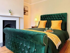 Un dormitorio con una cama tapizada de verde, una chimenea, una mesita de noche con una lámpara y un cuadro en la pared.