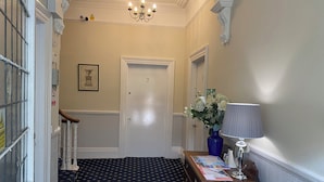 En hall med en dörr märkt med siffran ’2’, en kristallkrona, en blå vas med blommor och en lampa på ett träbord.