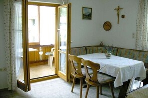 Ein Zimmer mit Esstisch, Stühlen, einer Bank, einem Fenster mit Vorhängen, einer Wanduhr und einem Kruzifix.