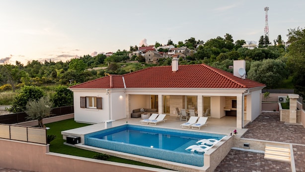Une maison moderne dotée d’une piscine, d’un patio avec des chaises longues et offrant une vue sur une colline où se trouvent d’autres maisons.