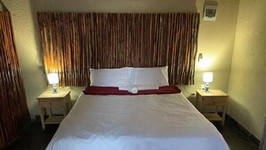 A neatly made bed with white bedding and a red accent, flanked by two bedside tables with lamps, in a room with a textured wall and a wooden headboard. - Xisakeni Lodge and Transfers  (Thulamahashe)