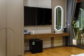A modern room with a flat-screen TV, a small refrigerator, a wooden desk, and a window with a view of plants.