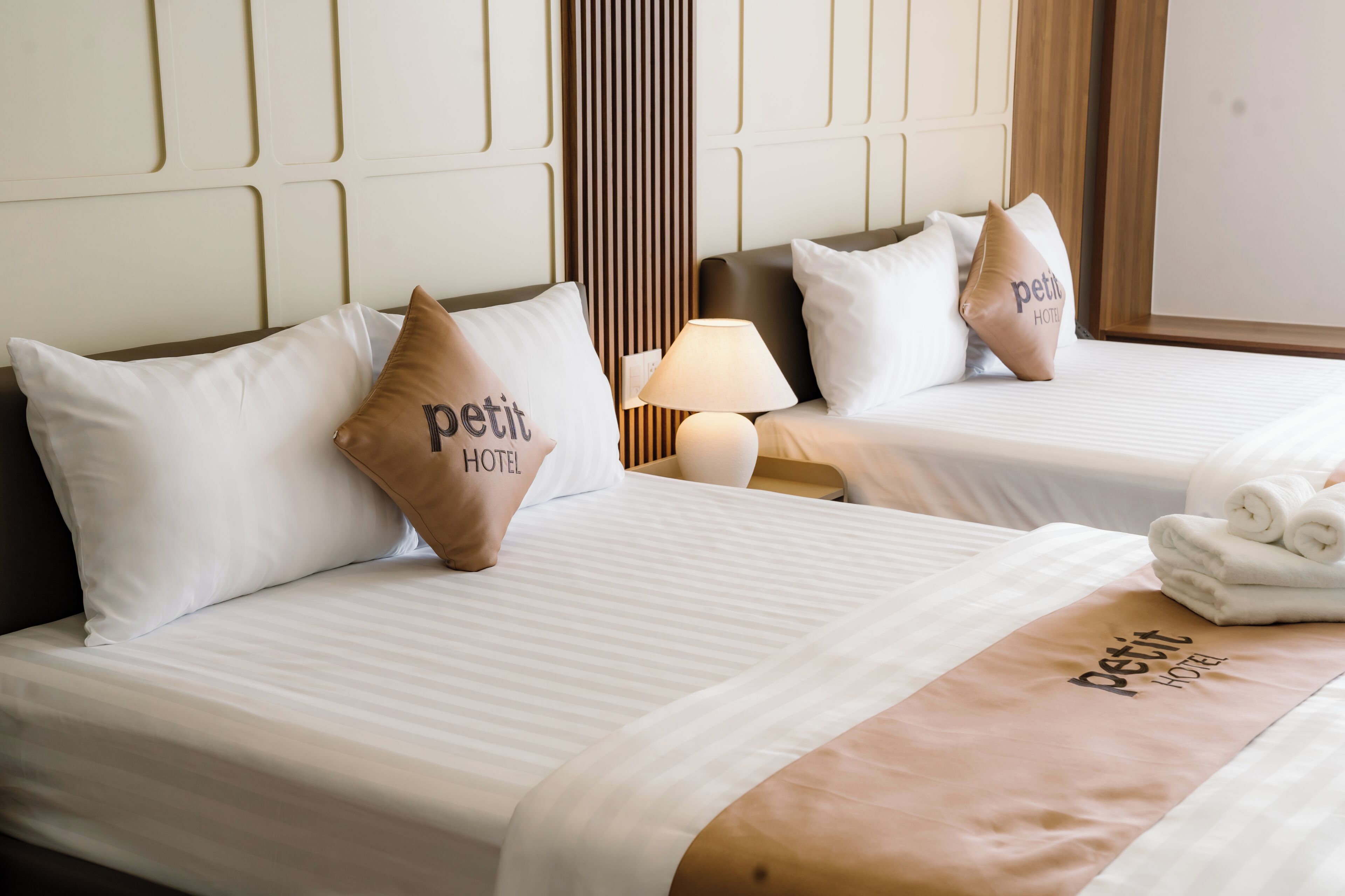 Two beds with white linens, a brown pillow with 'petit hotel' text, a bedside lamp, and a towel on the right bed.