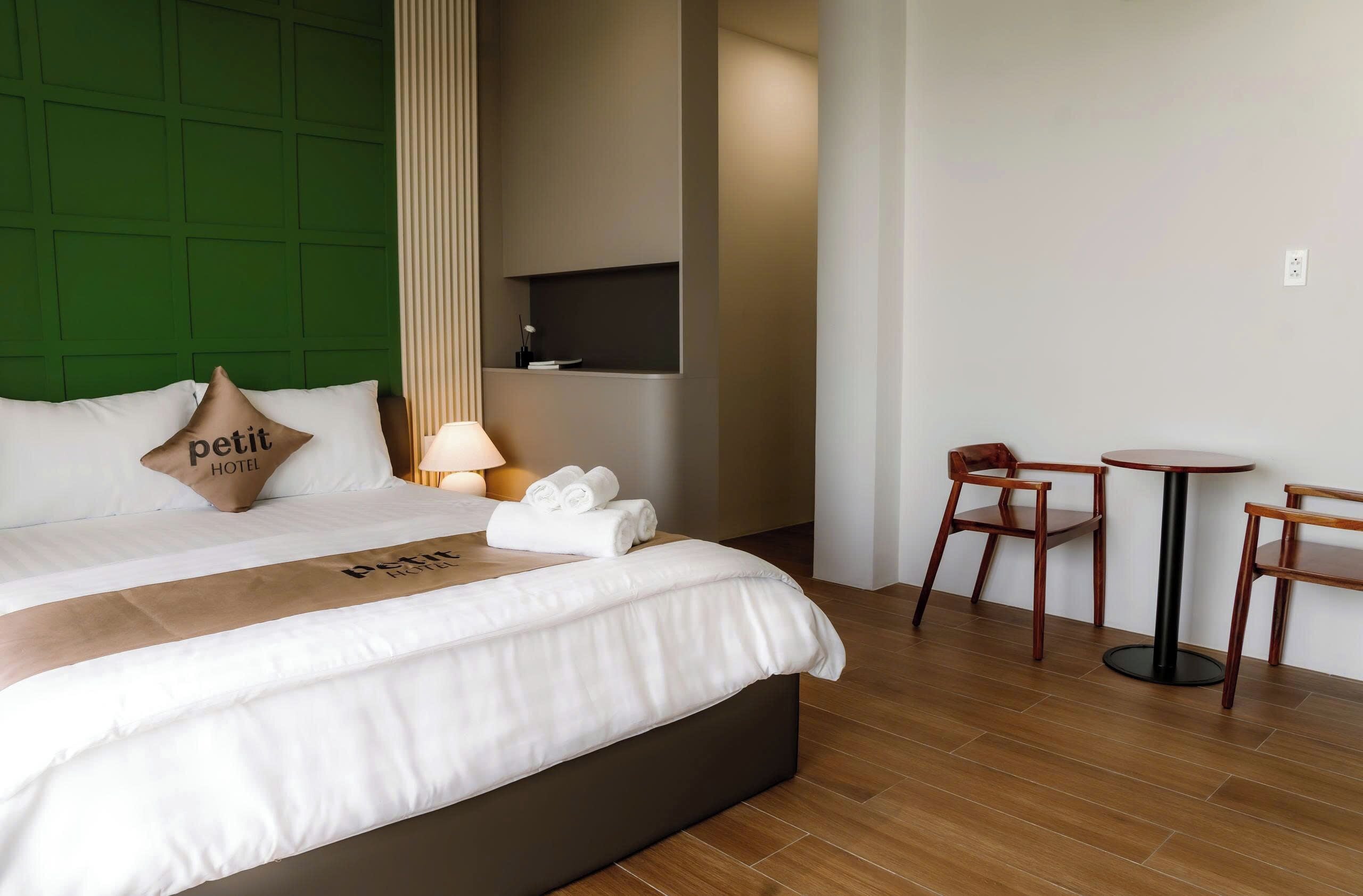 Una habitación de hotel con cama, cabecera verde, televisor montado en la pared, una pequeña mesa con sillas y una lámpara.