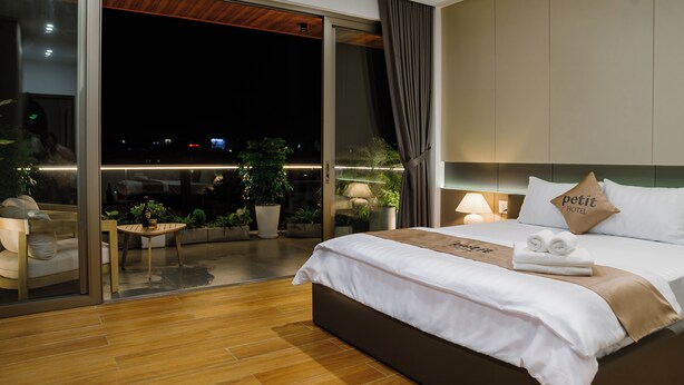 A modern hotel room with a large bed, a small table, a sofa, and a balcony with plants.