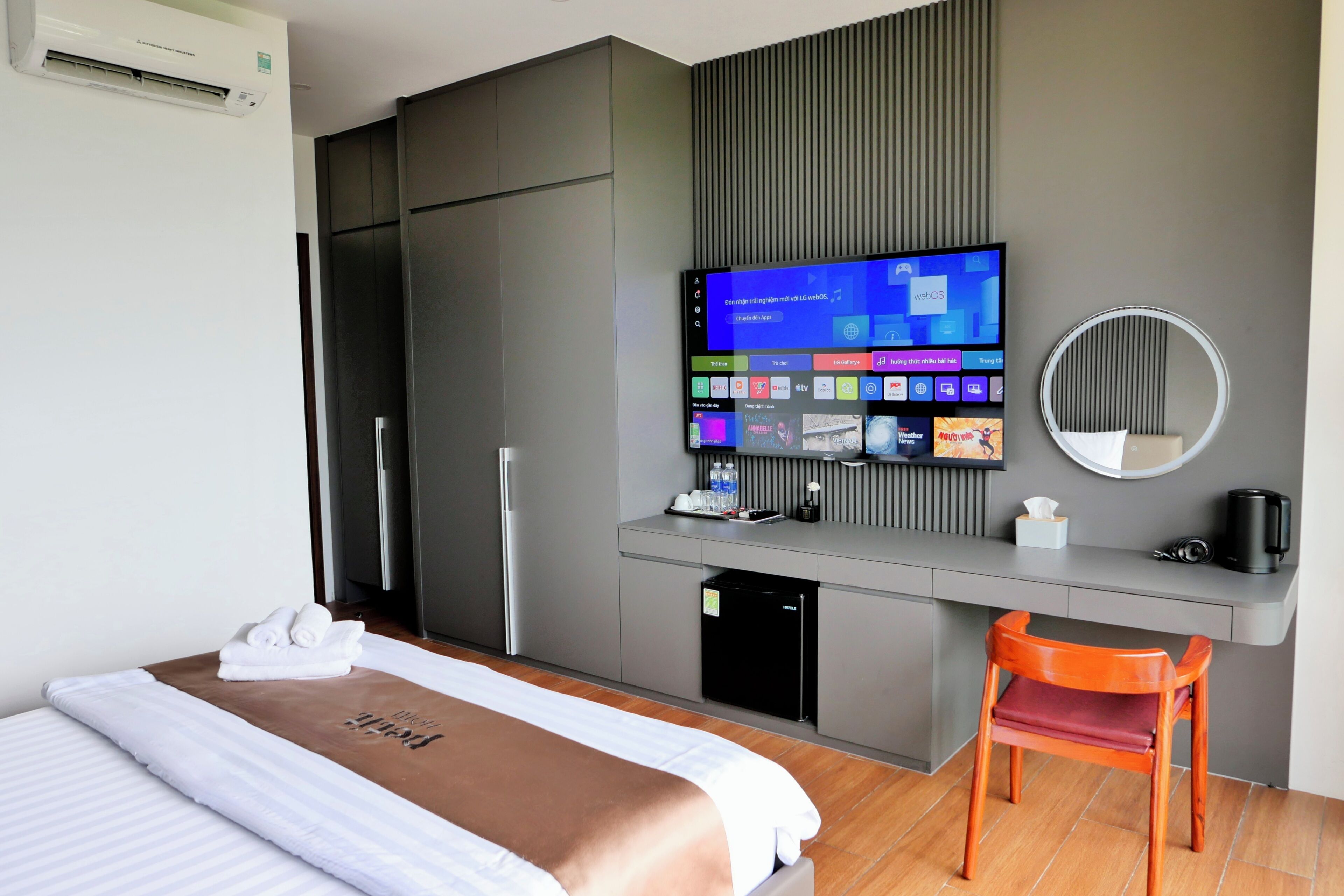 A modern hotel room with a bed, a desk with a flat-screen TV, a small refrigerator, and a chair.