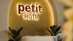 A modern hotel interior with a curved white sofa, a wall-mounted sign reading 'petit HOTEL,' and two potted plants.