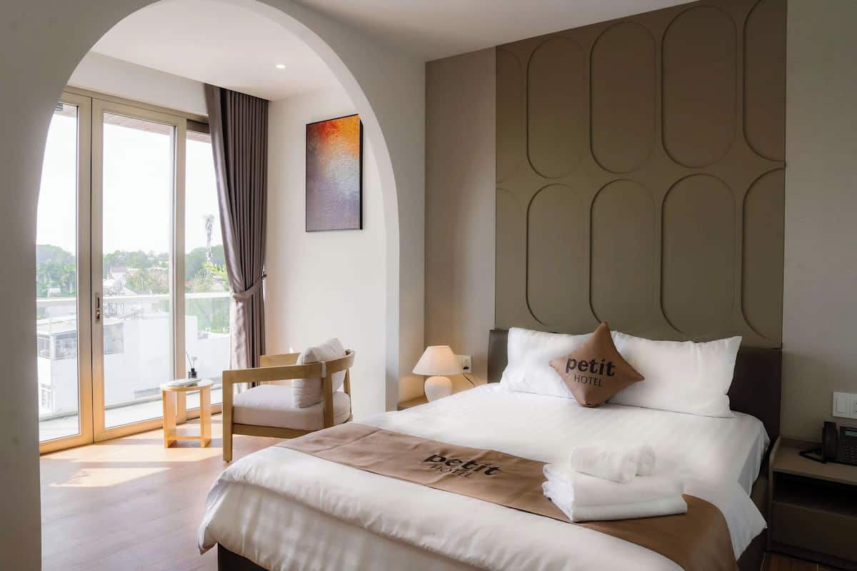 Une chambre d’hôtel avec un lit, une chaise, une table de chevet, une lampe et une vue sur l’extérieur grâce à une grande fenêtre.