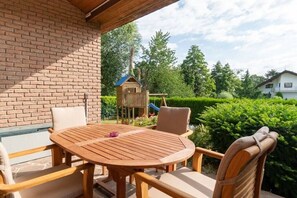 Une table et des chaises en bois, disposées dans un espace patio extérieur agrémenté d’un mur en briques, d’une maisonnette en bois et d’un jardin bien entretenu.