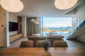A modern living room with a dining area, a flat-screen TV, a sofa, and a view of a pool and mountains.